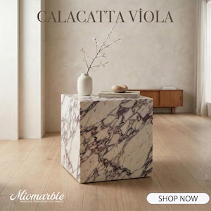 Pode incluir: Uma mesa lateral quadrada de mármore Calacatta Viola com um padrão branco e roxo. Um pequeno vaso branco com um raminho de flores, um livro e um objeto decorativo de madeira repousam no topo. O texto "CALACATTA VIOLA" está no topo.