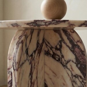 May include: A close-up of a marble table with a rounded base and a flat top. The marble has a white and purple veined pattern. A wooden sphere sits on the table's surface.