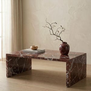 May include: A rectangular coffee table made of deep red marble with prominent white veining. A matching red marble vase containing a thin, bare, dark brown branch sits on the surface next to a small, smooth, light brown wooden bowl.