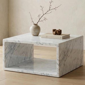 Könnte beinhalten: Ein quadratischer Couchtisch aus Marmor mit einem ausgehöhlten Mittelteil. Der Tisch hat graue Maserungen im weißen Marmor. Eine kleine Vase mit einem blühenden Zweig, ein Buch und eine Holzskulptur stehen oben.