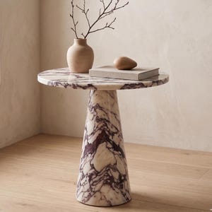 May include: A round marble side table with a white and purple veined pattern. A beige vase with a twig and a wooden decorative object sit on top of the table. The table has a conical base.
