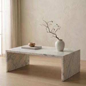 May include: A rectangular white marble coffee table with gray veining features a waterfall edge design. On the surface sits a small, smooth wooden bowl resting on a closed book, alongside a white ceramic vase containing a bare, dark brown branch.