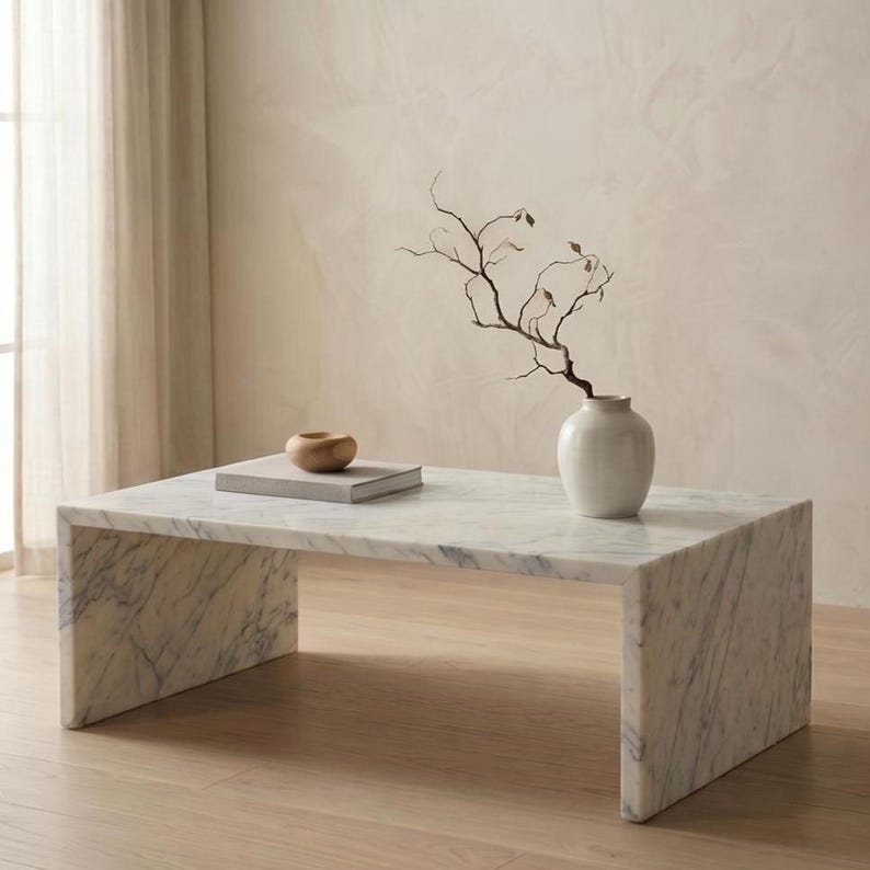 May include: A rectangular white marble coffee table with gray veining features a waterfall edge design. On the surface sits a small, smooth wooden bowl resting on a closed book, alongside a white ceramic vase containing a bare, dark brown branch.