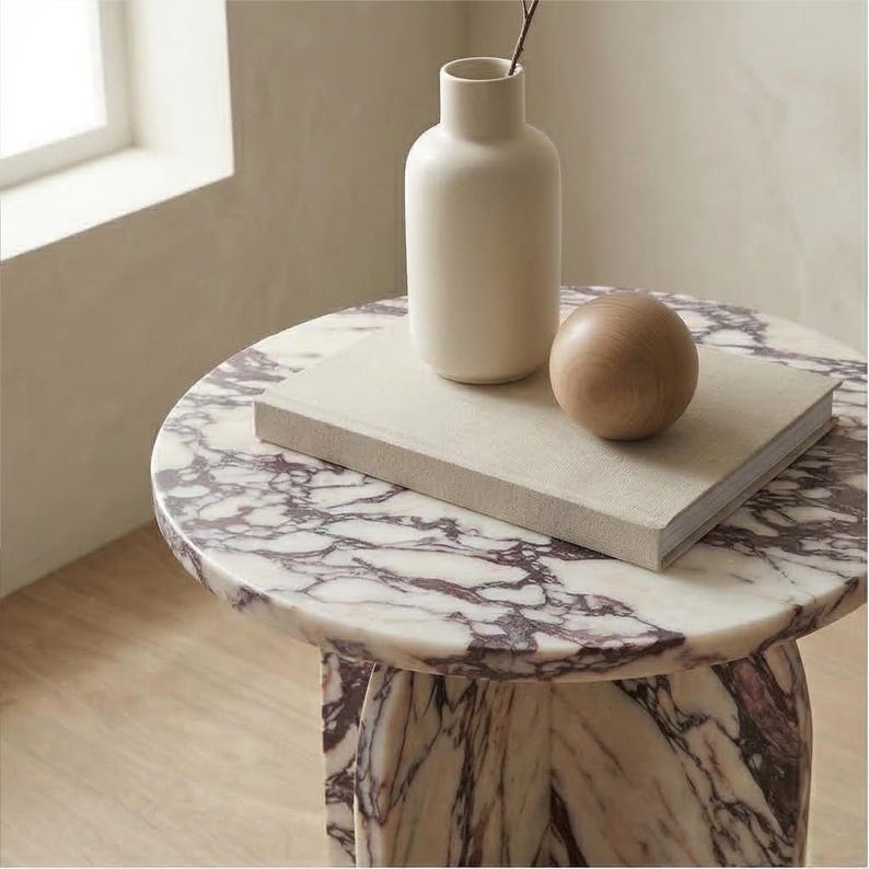 May include: A round marble side table with a white and purple veined pattern. A white vase, a wooden sphere, and a book rest on top. The table has a pedestal base and is placed on a wooden floor.