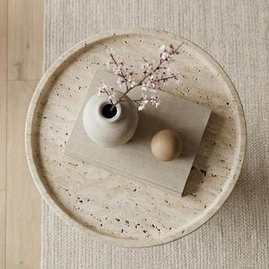 May include: A circular, beige tray with a speckled surface features a small, pale vase with pink blossom, a wooden sphere, and a rectangular, light-coloured book. The tray rests on a beige rug.