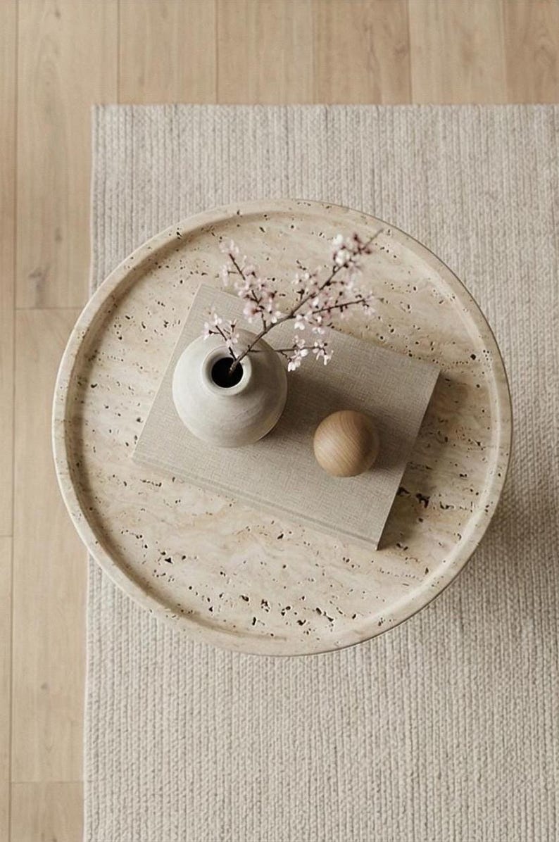 May include: A circular, beige tray with a speckled surface features a small, pale vase with pink blossom, a wooden sphere, and a rectangular, light-coloured book. The tray rests on a beige rug.