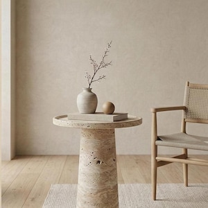 May include: A beige travertine side table with a round top and cylindrical base. A small vase with a sprig of flowers, a wooden sphere, and a book rest on the table. A woven chair sits nearby.
