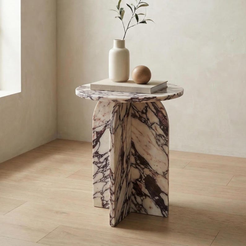May include: A round marble side table with a unique base. The table has a white and purple marble pattern. A white vase, a wooden sphere, and a stack of books sit on top. A sprig of greenery is in the vase.