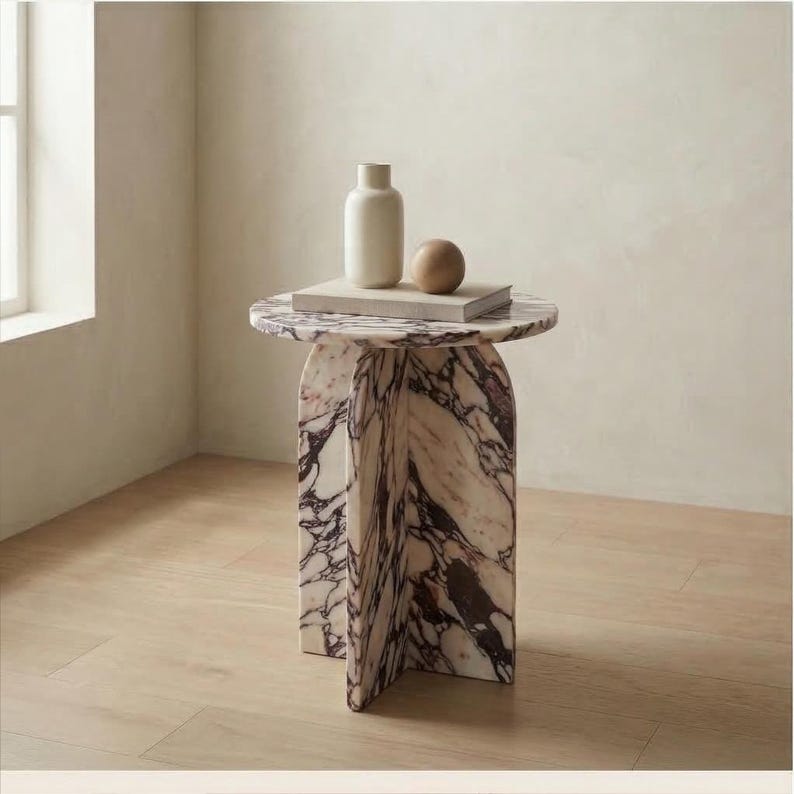 May include: A marble side table with a round top and arched legs. The table features a white and burgundy veined pattern. A white vase, a wooden sphere, and a book are on the table's surface. The table is approximately 20 inches in height.