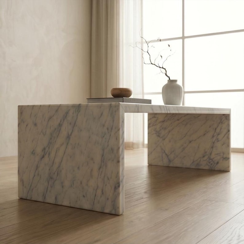 May include: A rectangular coffee table constructed from white marble with grey veining. The table features a waterfall edge design with two vertical side panels supporting a flat top surface. The surface is smooth and polished with a natural stone pattern.