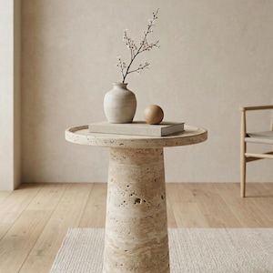 May include: A beige travertine side table with a round top and a cylindrical base. A small, off-white vase with a sprig of flowers, a wooden sphere, and a book rest on the table. The table is approximately 61 cm in height.