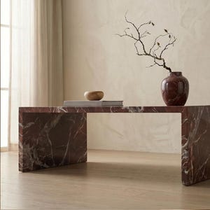 May include: A rectangular coffee table made of dark red marble with white veining. On top sits a dark red marble vase containing a thin, bare branch with small leaves, alongside a flat, light-colored book and a small, smooth wooden bowl.