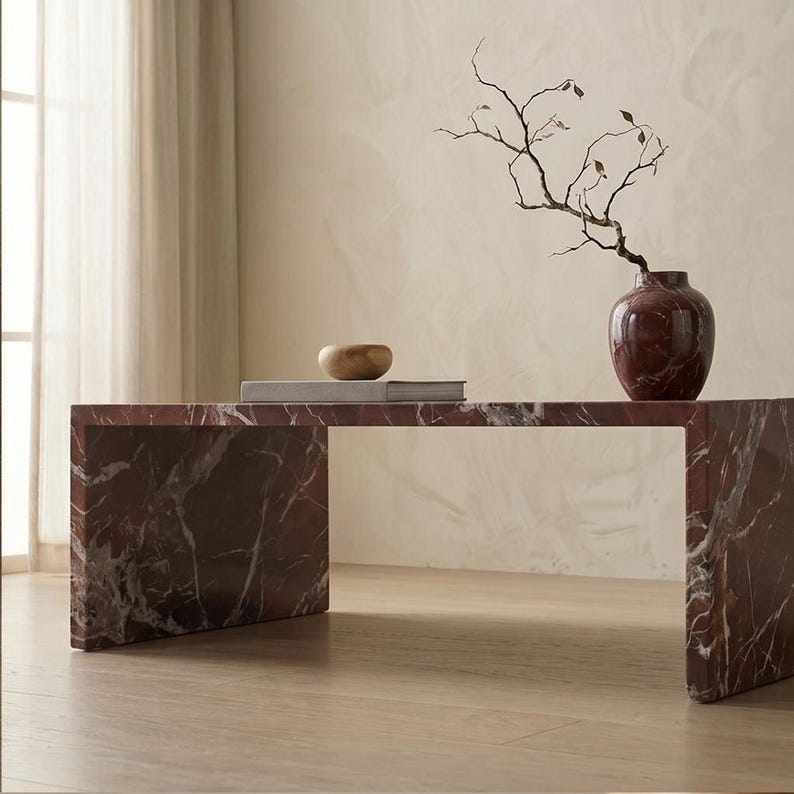 May include: A rectangular coffee table made of dark red marble with white veining. On top sits a dark red marble vase containing a thin, bare branch with small leaves, alongside a flat, light-colored book and a small, smooth wooden bowl.