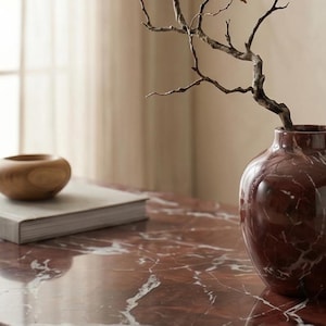 May include: A dark red marble vase with white veining holds a bare, twisted brown branch. Beside the vase sits a light-colored book and a small, smooth, rounded wooden bowl, all arranged on a polished dark red marble surface with white veining.