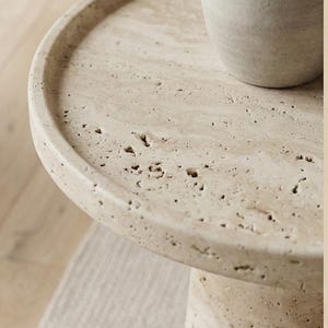 May include: A beige, round side table with a textured surface and a cylindrical base. A light-coloured vase sits on the table's surface. The table is made of a stone-like material and is approximately 50 cm in diameter.