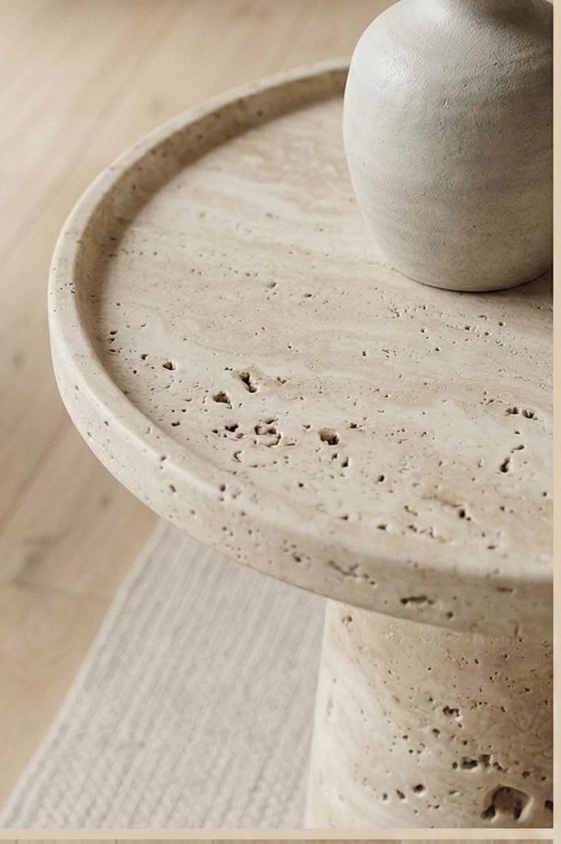 May include: A beige, round side table with a textured surface and a cylindrical base. A light-coloured vase sits on the table's surface. The table is made of a stone-like material and is approximately 50 cm in diameter.