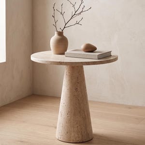 May include: A beige, round side table with a conical base. A small, beige vase with branches, a book, and a wooden decorative object sit on top. The table is made of a textured, stone-like material.