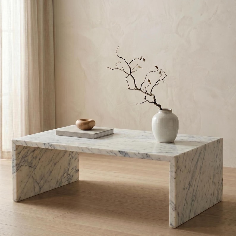 May include: A rectangular white marble coffee table with grey veining features a waterfall edge design. On the surface sits a small, smooth, round wooden bowl and a white ceramic vase containing a single, thin, dark, leafless branch.
