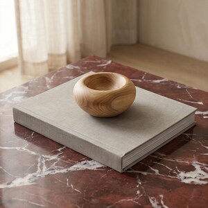 May include: A small, rounded wooden bowl with a smooth, light brown finish and visible natural wood grain patterns, resting on top of a square, light gray fabric-covered book with a thick spine and white pages.