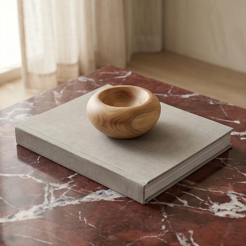 May include: A small, rounded wooden bowl with a smooth, light brown finish and visible natural wood grain patterns, resting on top of a square, light gray fabric-covered book with a thick spine and white pages.