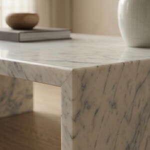 May include: A white marble coffee table with grey veining featuring a rectangular silhouette. On the surface sits a dark grey rectangular book and a small brown wooden bowl, with a portion of a white ceramic vase visible on the right side.