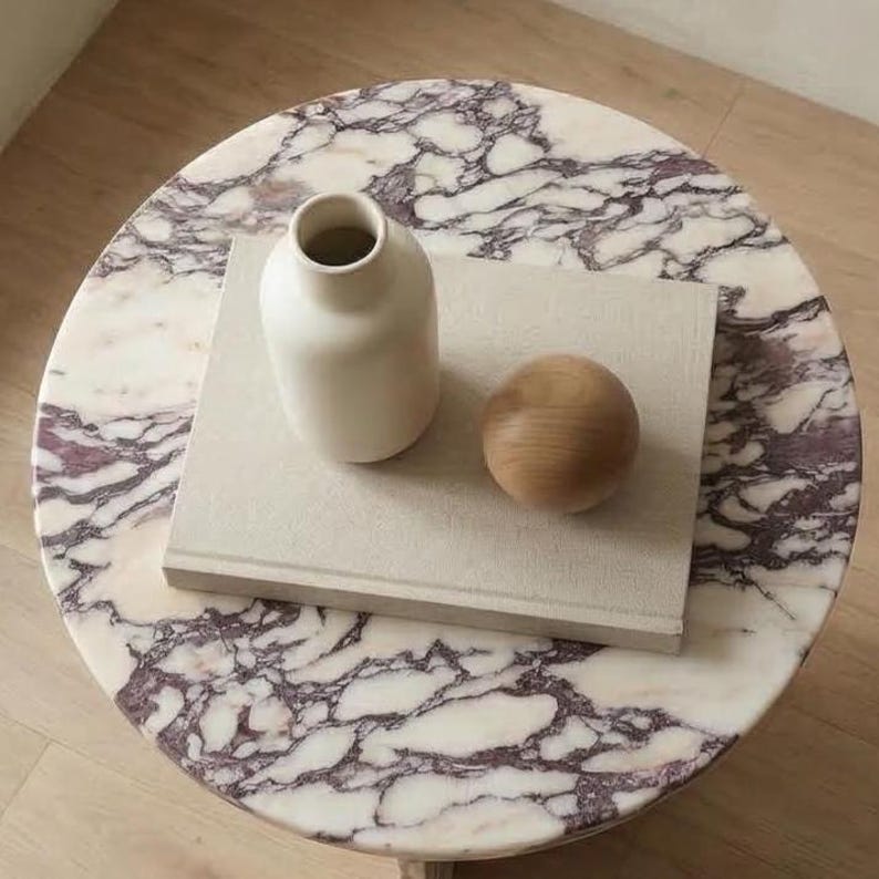 May include: A round marble table with a white and purple veined pattern. A white vase, a wooden sphere, and a beige book are on top of the table. The table is a decorative piece.
