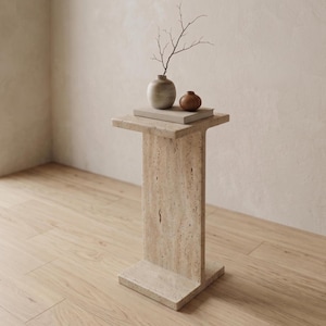 Peut inclure: Une table d'appoint en travertin beige avec un plateau et une base rectangulaires, dotée d'une colonne de support verticale. Sur la table se trouvent un livre rectangulaire, un vase en céramique arrondi clair, un petit bol en bois brun et une branche.