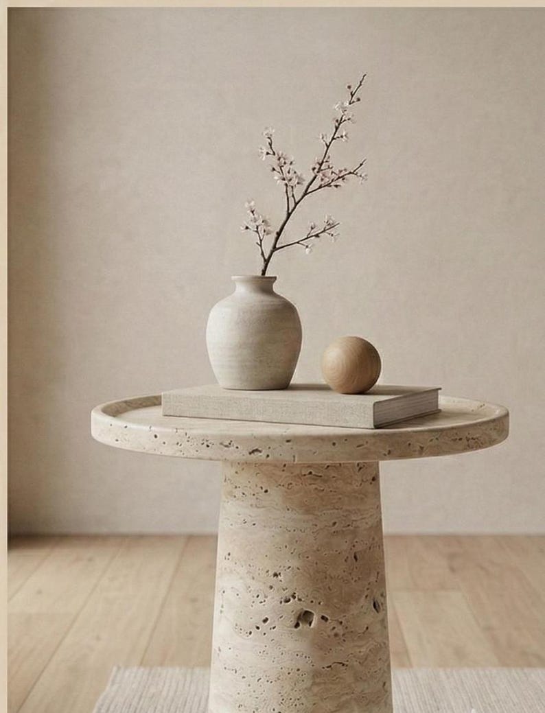 May include: A beige stone side table with a round top and a cylindrical base. A small, off-white vase with a flowering branch, a wooden sphere, and a book rest on the table. The table is set against a neutral background.