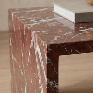 May include: A rectangular side table crafted from deep red marble with prominent white veining throughout the surface. The table features a clean, angular design with sharp edges and a polished finish, supporting a single grey book on its top surface.