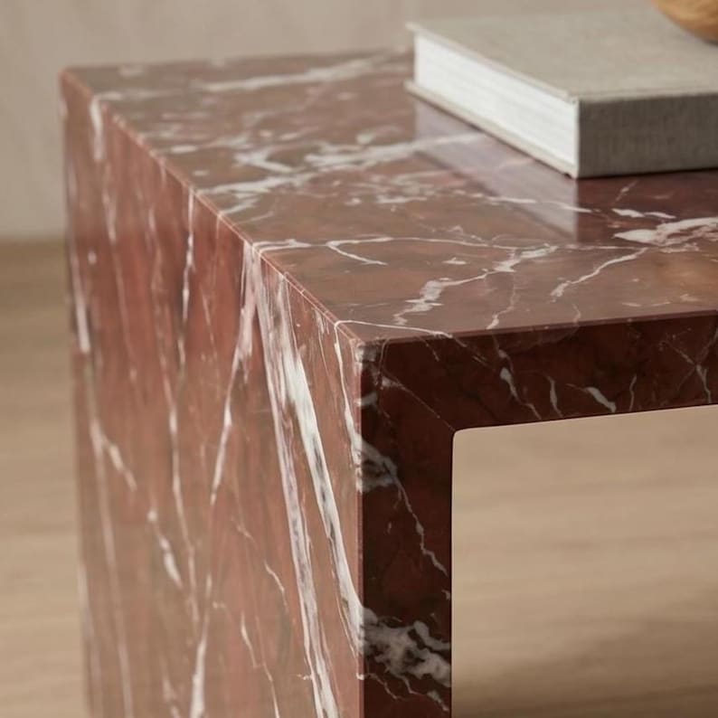May include: A rectangular side table crafted from deep red marble with prominent white veining throughout the surface. The table features a clean, angular design with sharp edges and a polished finish, supporting a single grey book on its top surface.
