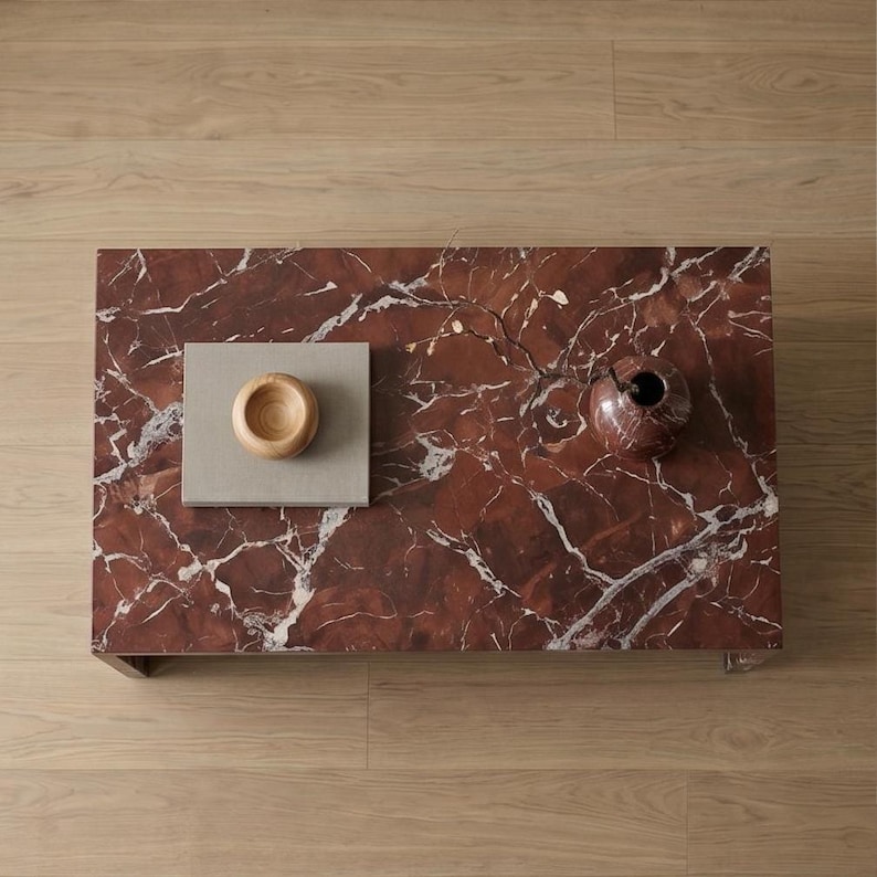 May include: A rectangular coffee table with a deep red marble top featuring prominent white veining. On the surface sits a square light gray book topped with a small, smooth, light-colored wooden bowl and a dark red marble vase with a thin branch.