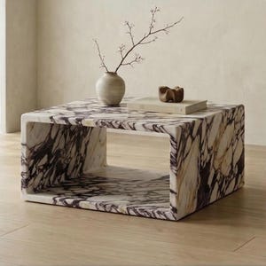 May include: A rectangular coffee table made of white marble with prominent dark purple and black veining. The table features a hollow center design. On top sits a light-colored ceramic vase containing a thin branch and a small beige book.