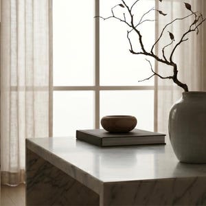 May include: A smooth, round wooden bowl sits atop a rectangular book with a dark cover. Beside the book, a light-colored ceramic vase holds a bare, branching twig with a few small, dried leaves attached to its thin, dark, winding stems.