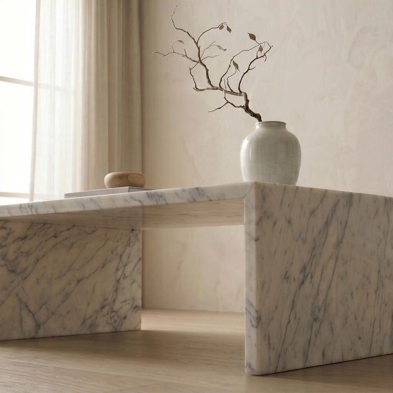 May include: A white marble coffee table with grey veining features a waterfall edge design. On the table sits a smooth, light-colored ceramic vase containing a single, bare, branching twig with a few small, dried brown leaves attached to it.