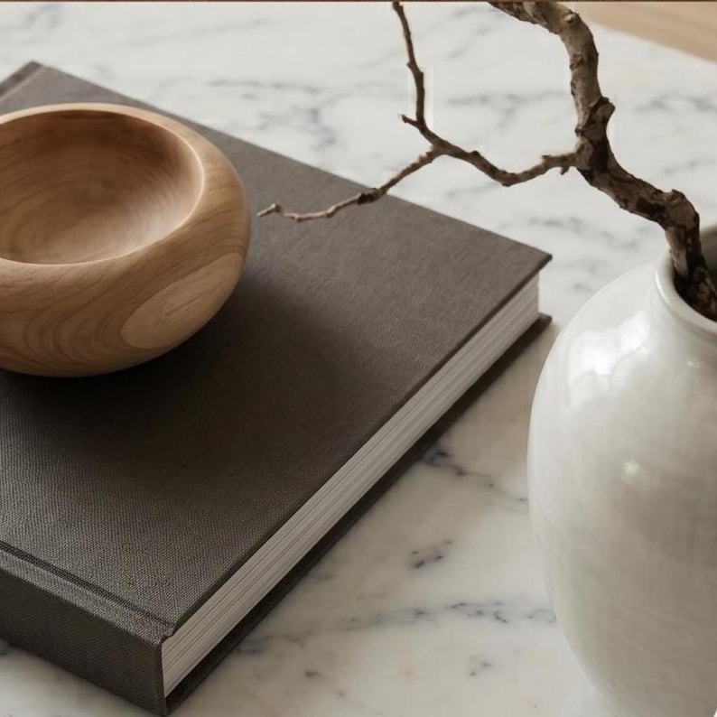 May include: A dark gray fabric-covered book positioned next to a small, smooth, light-colored wooden bowl. To the right, a white ceramic vase holds a thin, bare, brown branch. The objects are arranged together on a white marble surface.