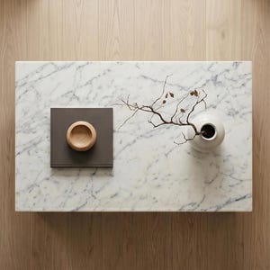 May include: A rectangular white marble coffee table with grey veining. On the surface sits a square dark grey book, a small light brown wooden bowl, and a white ceramic vase containing a thin, bare branch with a few small brown leaves.