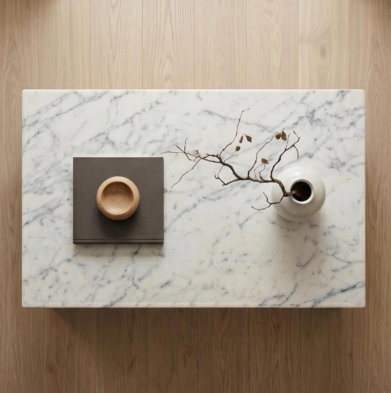 May include: A rectangular white marble coffee table with grey veining. On the surface sits a square dark grey book, a small light brown wooden bowl, and a white ceramic vase containing a thin, bare branch with a few small brown leaves.