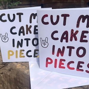 May include: Two white greeting cards featuring the black text "CUT MY CAKE INTO PIECES" and a hand gesture icon. The front card has red and black glittery lettering, while the back card features black and gold lettering. Both cards are folded and upright.