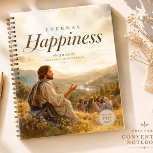 Può includere: Un quaderno a spirale con le parole "Eternal Happiness" e "2026 Convention Notebook" sulla copertina. La copertina presenta un dipinto di un uomo che parla a un gruppo di persone in un paesaggio. Una penna dorata è appoggiata accanto al quaderno.