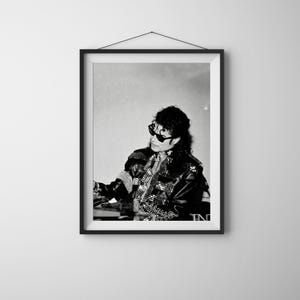 May include: Black and white framed print of a person wearing sunglasses and a studded jacket. The person is seated and appears to be performing. The image is a portrait and is displayed on a white wall.