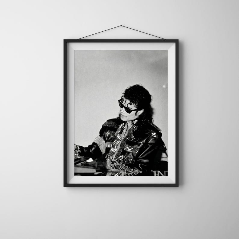May include: Black and white framed print of a person wearing sunglasses and a studded jacket. The person is seated and appears to be performing. The image is a portrait and is displayed on a white wall.
