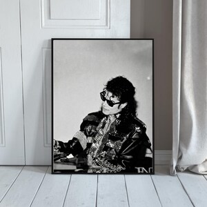 May include: Black and white framed print featuring a portrait of a person wearing sunglasses and a black jacket with silver embellishments. The image is set against a neutral background, with a white door and wooden floor visible.