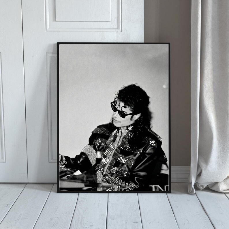May include: Black and white framed print featuring a portrait of a person wearing sunglasses and a black jacket with silver embellishments. The image is set against a neutral background, with a white door and wooden floor visible.