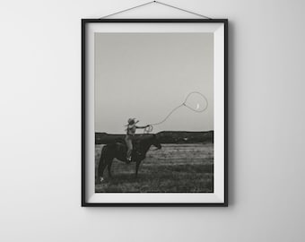 May include: A black and white framed wall art print depicting a person on horseback in a field, swinging a lasso into the air. The scene is set against a vast, open sky with a crescent moon visible within the loop of the rope, captured in a monochromatic style.