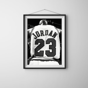 May include: Black and white framed print of a basketball jersey. The jersey is white with the name "JORDAN" arched across the top and the number "23" in large black numerals below. The image is a close-up of the back of the jersey.