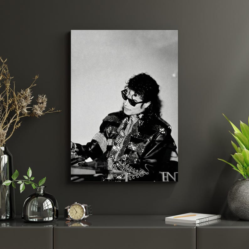 May include: Black and white framed print of Michael Jackson wearing sunglasses and a studded jacket. The image captures a moment of the artist seated, with a focused expression. The print is displayed on a dark gray wall, adding a touch of elegance.