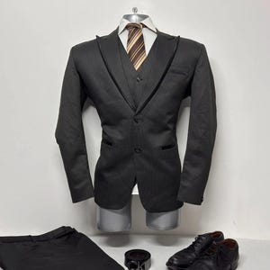 May include: A dark gray three-piece suit displayed on a mannequin, including a blazer, vest, and trousers. A striped tie, black belt, and black leather shoes complete the ensemble. The suit is displayed against a white background.