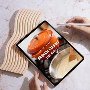 以下が含まれることがあります： タブレットには、「French Cuisine Easy Basics」というタイトルの料理本の表紙とオレンジ色の鍋が表示されています。木製の波型のオブジェクトとスタイラスも表示されています。背景は大理石の表面です。
