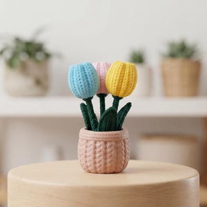 May include: A small, decorative arrangement of three crocheted tulips in a pink pot. The tulips are blue, pink, and yellow, with green stems and leaves. The pot has a textured, woven appearance. The arrangement sits on a light-colored wooden surface.