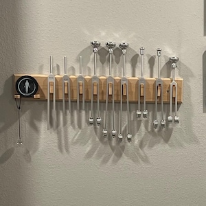 May include: A wall-mounted display featuring a collection of tuning forks, a macrame shelf with a clock, and two framed certificates. The tuning forks are silver and mounted on a wooden rack. The macrame shelf has a wooden shelf and tassels.